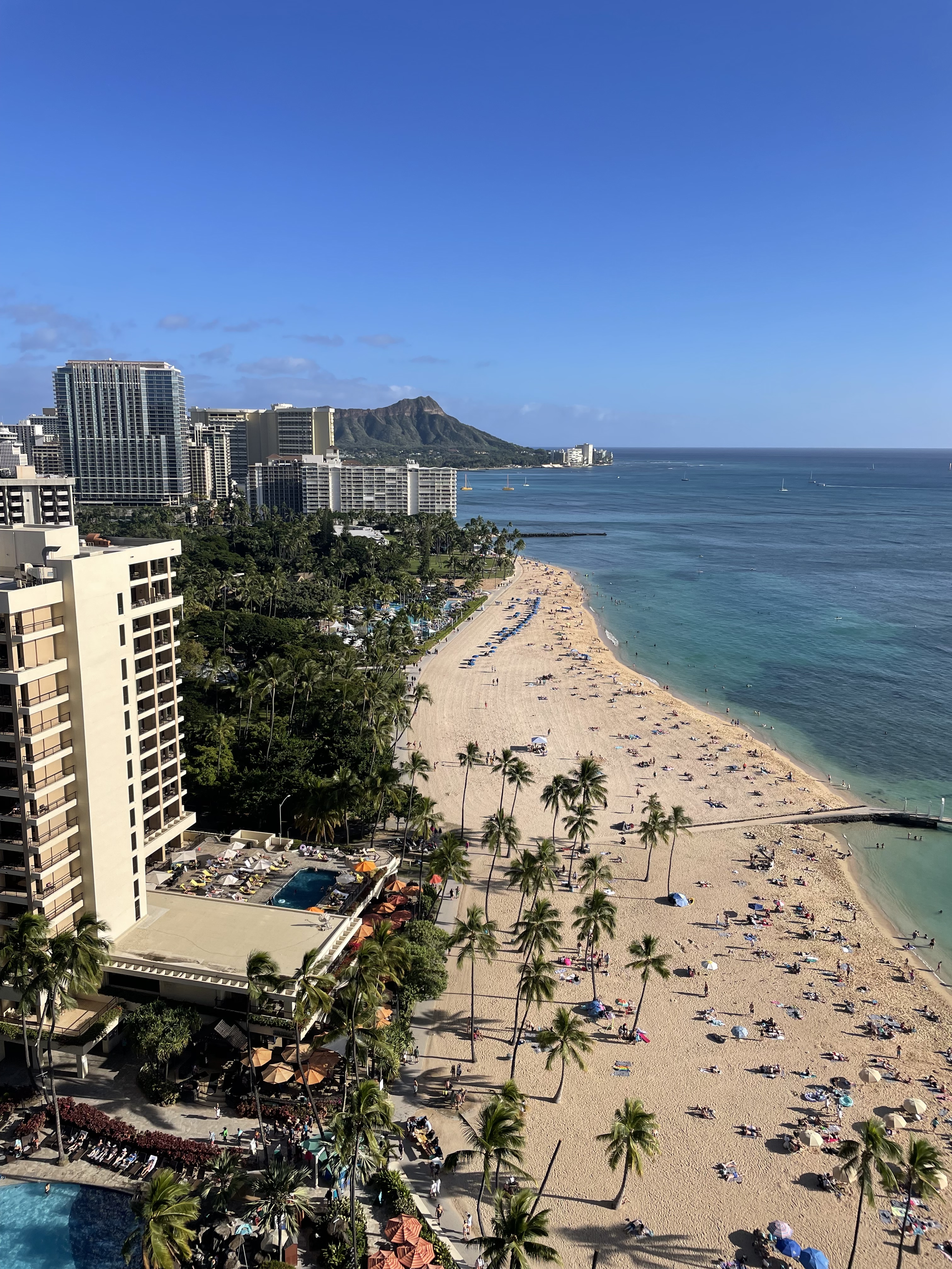Aerial view of a curving city beach lined with hotel towers and palms, with a headland beyond.